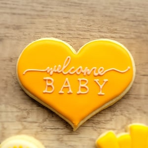 May include: A yellow heart-shaped cookie decorated with pink icing and the words "welcome baby".