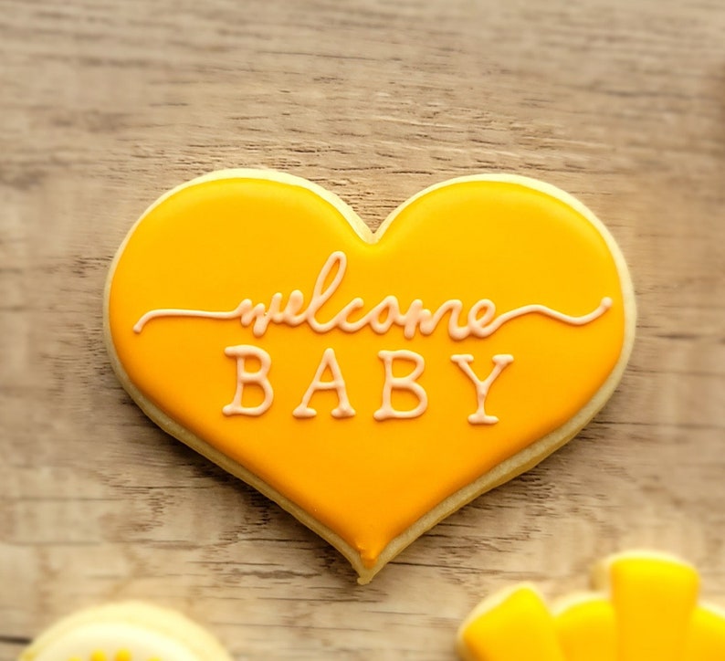 May include: A yellow heart-shaped cookie decorated with pink icing and the words "welcome baby".