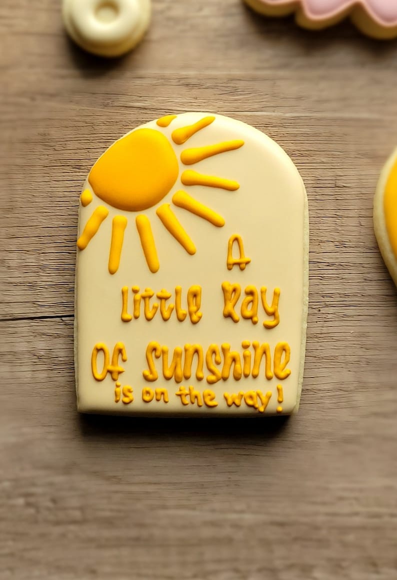 May include: A yellow and white cookie decorated with a yellow sun and the text "A little ray of sunshine is on the way!"