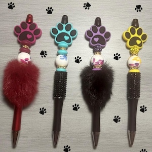 May include: Four decorative pens with paw print toppers in pink, teal, purple, and yellow. Each pen has a different colored fluffy pom-pom and a beaded design. The pen barrels are black and silver.