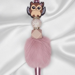 May include: A pink pen with a decorative owl topper. The owl is brown and tan with black accents and a small pink bow. The pen has a pink fur accent and a geometric bead.