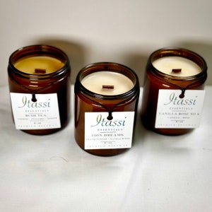 May include: Three amber glass jars with wooden lids and labels. The labels are white with black text and say "Itassi Essentials" and the name of the scent. The scents are "Bush Tea", "Zion Dreams", and "Vanilla Rose No. 6".
