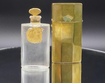 May include: A clear glass perfume bottle with a gold-colored emblem and stopper, next to a gold-colored rectangular case with a green and gold design. The bottle and case are on a reflective surface.