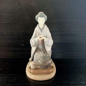 Può includere: Una statuetta in porcellana raffigurante una persona in kimono grigio chiaro inginocchiata mentre tiene in mano una tazzina. La figura è posizionata dietro un vassoio con una teiera grigio scuro e una tazzina bianca, su una base circolare.