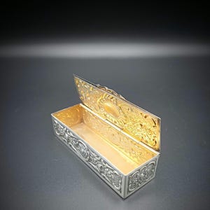 May include: An open rectangular silver box with intricate floral and geometric patterns. The interior is gold-colored, and the lid is slightly ajar, revealing the gold interior and ornate designs. The box is set against a dark background.