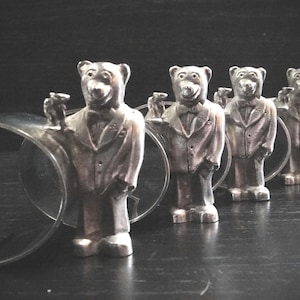 Peut inclure: Un ensemble de sept figurines d'ours argentées, chacune portant un costume et tenant un petit objet. Les ours sont disposés en ligne, le plus grand ours en tête et le plus petit ours à l'arrière. Les ours sont tous debout sur une surface noire.