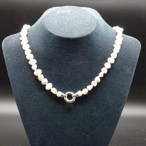 May include: A white pearl necklace with a silver clasp is displayed on a black jewelry bust. The pearls are small and irregularly shaped. The clasp is a circular design. The background is black.