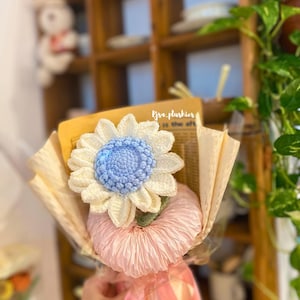 May include: A crocheted sunflower bouquet with a blue center and white petals. The bouquet is wrapped in brown paper and tied with a pink ribbon. The text "lysa_plushies" is visible in the background.