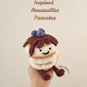 May include: A crocheted pancake plush toy inspired by Jellycat. The plush toy is brown and white with a purple blueberry on top. The toy has a smiling face and two brown legs.