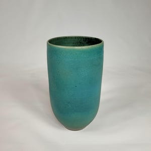 Könnte beinhalten: Eine hohe, zylindrische Keramikvase mit strukturierter Oberfläche. Die Vase hat einen Farbverlauf aus Türkis- und Türkistönen mit dunklerem Rand. Die Gesamtform ist leicht konisch, und der Boden hat einen helleren Farbton derselben Farbfamilie.