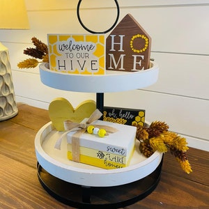 May include: A two-tiered decorative tray with bee-themed decor. The top tier features a sign that says "Welcome to our Hive" and a wooden house with "HOME" on it. The bottom tier has a stack of books that say "sweet like honey".