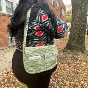 May include: A green crocheted shoulder bag with a white lace trim. The bag has a pocket on the front and is worn over the shoulder.
