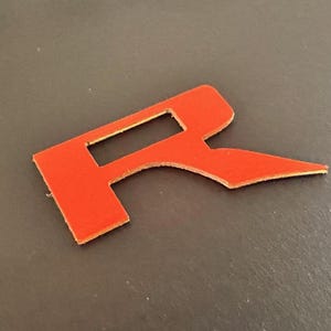 Puede incluir: Corte de letra R de cuero rojo. La letra est&aacute; estilizada con un dise&ntilde;o &uacute;nico.