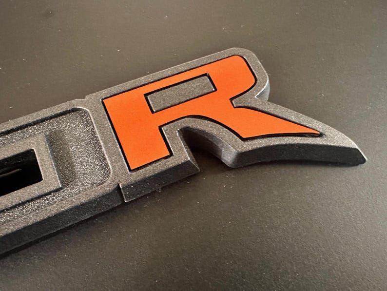 Puede incluir: Primer plano de un emblema de pl&aacute;stico negro "R" con un acento naranja. El emblema forma parte de una palabra m&aacute;s grande, probablemente "Raptor".