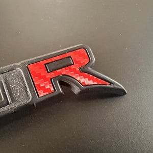 Puede incluir: Un emblema de pl&aacute;stico negro con una "R" roja de estilo fibra de carbono en el centro. El emblema est&aacute; dise&ntilde;ado para ser fijado a un veh&iacute;culo.