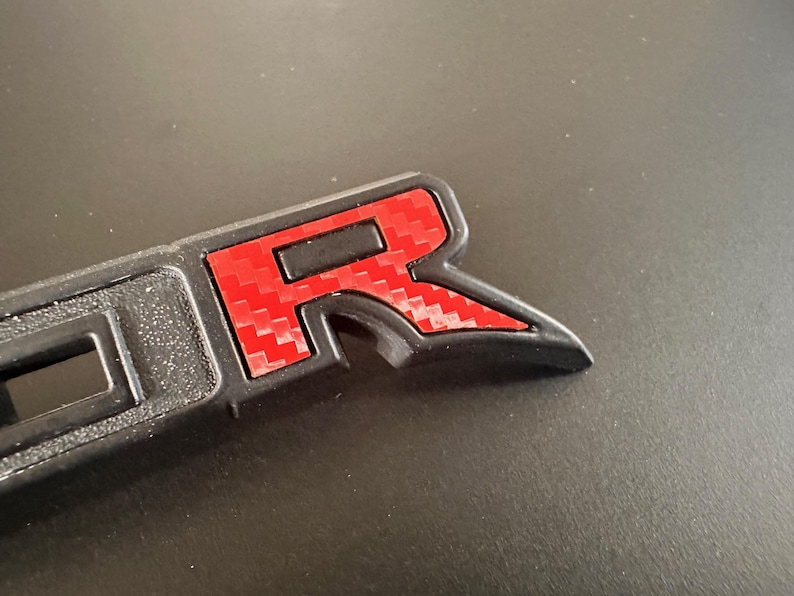 Puede incluir: Un emblema de pl&aacute;stico negro con una "R" roja de estilo fibra de carbono en el centro. El emblema est&aacute; dise&ntilde;ado para ser fijado a un veh&iacute;culo.
