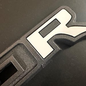 Puede incluir: Primer plano de un emblema de pl&aacute;stico negro con una "R" blanca en una fuente estilizada. El emblema tiene una superficie texturizada en relieve.