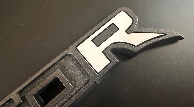 Puede incluir: Primer plano de un emblema de pl&aacute;stico negro con una "R" blanca en una fuente estilizada. El emblema tiene una superficie texturizada en relieve.