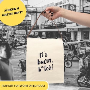 May include: A white canvas tote bag with brown leather straps. The bag has black text that reads "It's baon. b*tch!"  The bag is being held by a hand in front of a black and white photo of a street scene.