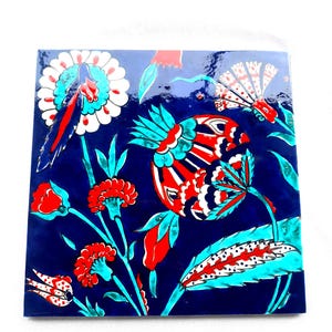 Könnte beinhalten: Quadratische Keramikfliese mit glänzendem Finish, mit einem lebendigen Blumendesign. Das Kunstwerk zeigt rote, weiße und türkisfarbene Blumen und Blätter vor einem tiefblauen Hintergrund. Die Fliese ist ein dekoratives Stück.