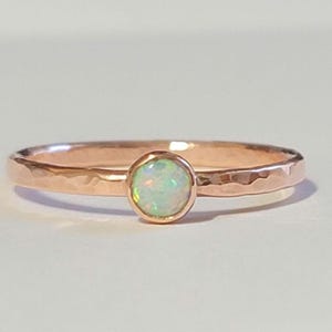 May include: A rose gold ring with a hammered band and a round, iridescent opal gemstone. The opal has a milky white base with flashes of green and blue. The ring is set against a plain white background.