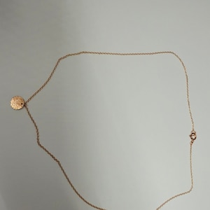 May include: A delicate gold-colored necklace with a small, textured circular pendant. The chain is fine and the clasp is visible. The necklace is displayed on a light-colored surface.