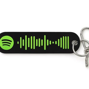 Könnte beinhalten: Schwarzer Schlüsselanhänger mit grünem Spotify-Logo und Wellenmuster. Er hat einen silberfarbenen Verschluss zum Befestigen an Schlüsseln oder Taschen. Das Spotify-Logo befindet sich auf der linken Seite des Schlüsselanhängers.
