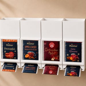Könnte beinhalten: Ein weißer, an der Wand befestigter Teebeutel-Organizer mit vier Fächern. Jedes Fach enthält Teebeutel mit verschiedenen Geschmacksrichtungen, darunter "Bratapfel" und "Zimt-Pflaume". Die Teebeutelverpackung zeigt Illustrationen von Tassen und Früchten.