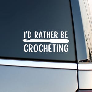 May include: White vinyl decal with the text "I'd rather be crocheting" and a crochet hook graphic. The decal is displayed on a car window, suggesting it's a car accessory for craft enthusiasts.
