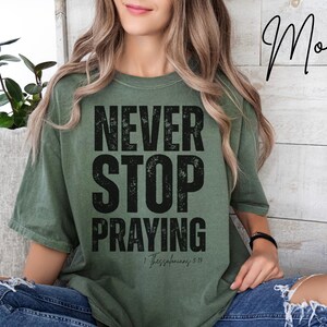 Puede incluir: Camiseta verde oliva con las palabras "NEVER STOP PRAYING" en letras negras grandes y desgastadas. El texto "1 Tesalonicenses 5:17" está debajo. La persona lleva una camiseta con pelo largo y ondulado.