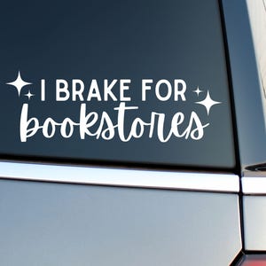 May include: White vinyl decal with the text "I Brake for Bookstores" in a whimsical font, accented with starburst graphics. The sticker is applied to the rear window of a vehicle.