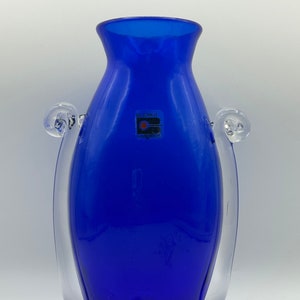 Può includere: Un vaso in vetro blu cobalto con due manici in vetro trasparente. Il vaso ha un'etichetta che recita "Blenko Hand Made".
