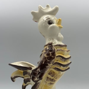 Puede incluir: Una figura de gallo de cerámica pintada en marrón, amarillo y blanco. El gallo tiene una cabeza blanca con un pico amarillo y ojos negros. El gallo está de pie sobre una base verde con una cola marrón.