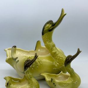 Op de afbeelding: Een set van drie groene keramische zwanen potten. De grootste zwaan heeft zijn nek omhoog gebogen en de twee kleinere zwanen hebben hun nek naar beneden gebogen. De zwanen zijn allemaal geglazuurd in een lichtgroene kleur met donkergroene accenten.