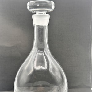 Può includere: Una caraffa di vetro trasparente con tappo. La caraffa ha un corpo bulboso e un collo lungo e sottile. Il tappo è rettangolare e si adatta perfettamente al collo. Il fondo della caraffa presenta un testo inciso.