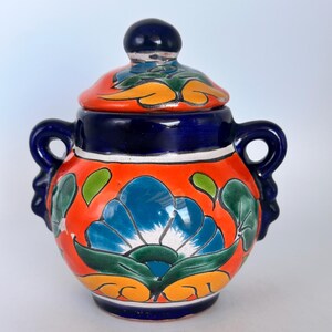 Op de afbeelding: Handbeschilderde keramische pot met een deksel. De pot is rond met twee handvatten en is versierd met een blauwe bloem en groene bladeren op een oranje achtergrond. Het deksel is oranje met blauwe en groene accenten.