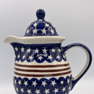 Puede incluir: Una jarra de cerámica azul y blanca con tapa, decorada con un patrón de estrellas blancas sobre fondo azul y rayas rojas. La jarra tiene un cuerpo redondeado y un asa.