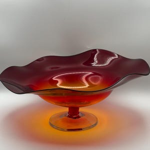 May include: A vibrant, red-orange glass bowl with a ruffled rim. The bowl sits on a short, round stem and base. The glass has a glossy finish, reflecting light and creating a gradient of colors.
