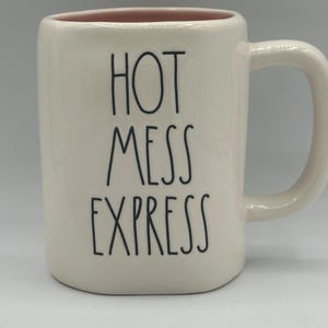 Puede incluir: Taza de cerámica color crema con las palabras "HOT MESS EXPRESS" en letras negras. El interior de la taza es rosa. La taza tiene un asa curvada.