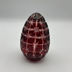 Puede incluir: Un objeto decorativo en forma de huevo, de color rojo rubí. La superficie está intrincadamente tallada con un patrón geométrico de líneas que se cruzan, creando un efecto de explosión estelar. El objeto está hecho de un material transparente, probablemente vidrio o cristal.