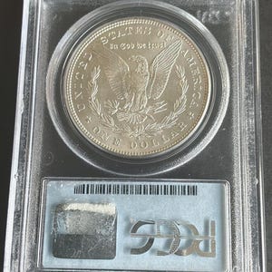 May include: A silver One Dollar coin, in a clear protective case. The coin shows an eagle with spread wings and the inscriptions "United States of America" and "In God we trust". The case has a label with a barcode and the letters "PCGS".