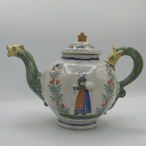 May include: A white ceramic teapot with green dragon-shaped handles and a floral design. The teapot has a lid with a knob in the shape of a star. The teapot is decorated with a painted scene of a woman in a blue dress and orange apron.