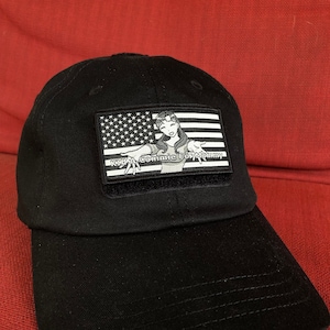 Könnte beinhalten: Eine schwarze Baseballkappe mit einem Patch, das eine Comicfigur einer Frau vor einem amerikanischen Flaggenhintergrund zeigt und den Text "K.I.K.A. Comic For Mommy" trägt.