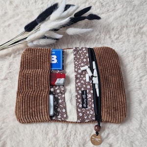 Könnte beinhalten: Ein braunes Kord-Etui mit Reißverschluss und einem runden Holzanhänger. Das Innere ist mit braunem Stoff mit weißem Blumenmuster und einem cremefarbenen Bereich ausgekleidet. Es enthält eine blaue Karte, ein rotes Feuerzeug und weiße Stäbchen.