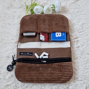Könnte beinhalten: Eine braune Cordtasche mit drei offenen Fächern, die ein silbernes Metallgehäuse, ein rotes Feuerzeug und eine blaue Karte enthalten. Darunter befindet sich ein Reißverschlussfach mit kleinen weißen zylindrischen Objekten. Etikett Betty Sue.