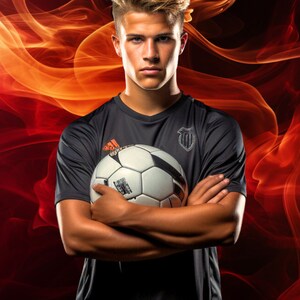 May include: A young man in a black soccer jersey with a white soccer ball in his arms. He is standing in front of a red and orange smoke background.