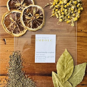 May include: An overhead view of dried lemon slices, chamomile flowers, herbs, and bay leaves on a wooden surface. A card reads "plant magick IMBOLC intentionally crafted for inner awakening, growth & new life by MELISSA SHAW."