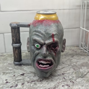 May include: A novelty beer mug shaped like a zombie head. The mug is gray with red accents, a green eye, and a handle. A beer can is inserted into the top of the head.