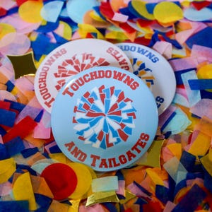 Puede incluir: Tres botones redondos con las palabras "Touchdowns and Tailgates" sobre un fondo de confeti colorido. Los botones son de color azul claro y blanco, con detalles en rojo y azul. Los botones son probablemente para un evento temático de fútbol americano.