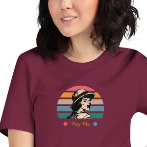 May include: A maroon short-sleeved t-shirt with a retro-style graphic of a woman wearing a hat in front of a sunset. The graphic includes the text "Pay Me".